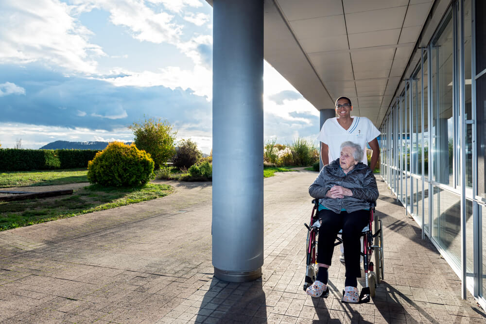 Un soignant avec une résidente en fauteuil roulant dans le parc de Mon Repos.
